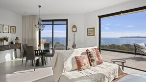 Een ruime woonkamer met een groot raam met uitzicht op zee, een eethoek met een tafel en stoelen, en een gezellige zithoek met een bank en kussens.