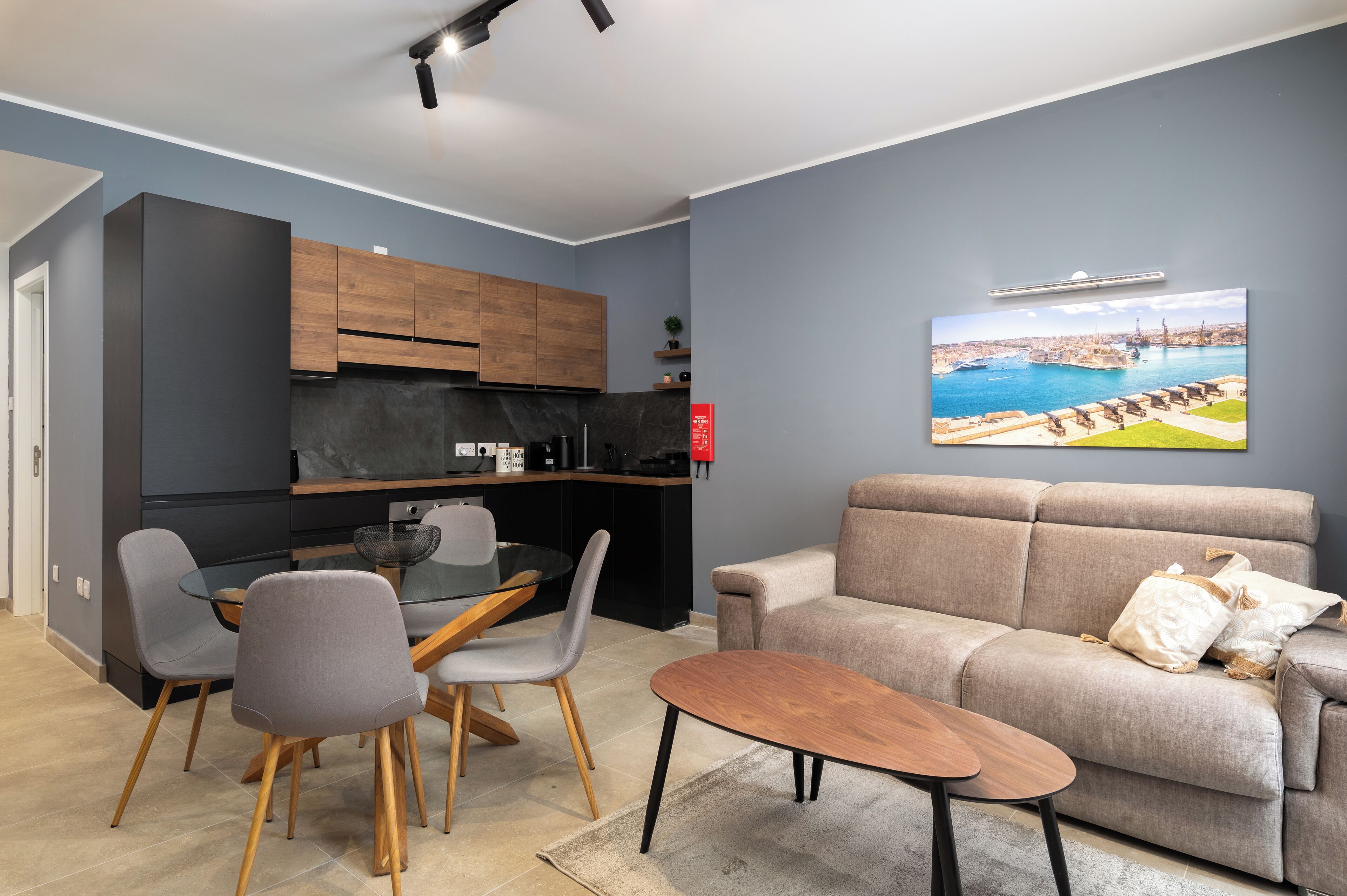 Una sala de estar moderna con un sofá gris, una mesa de comedor de vidrio con sillas grises, y una cocina con armarios oscuros y encimera negra.