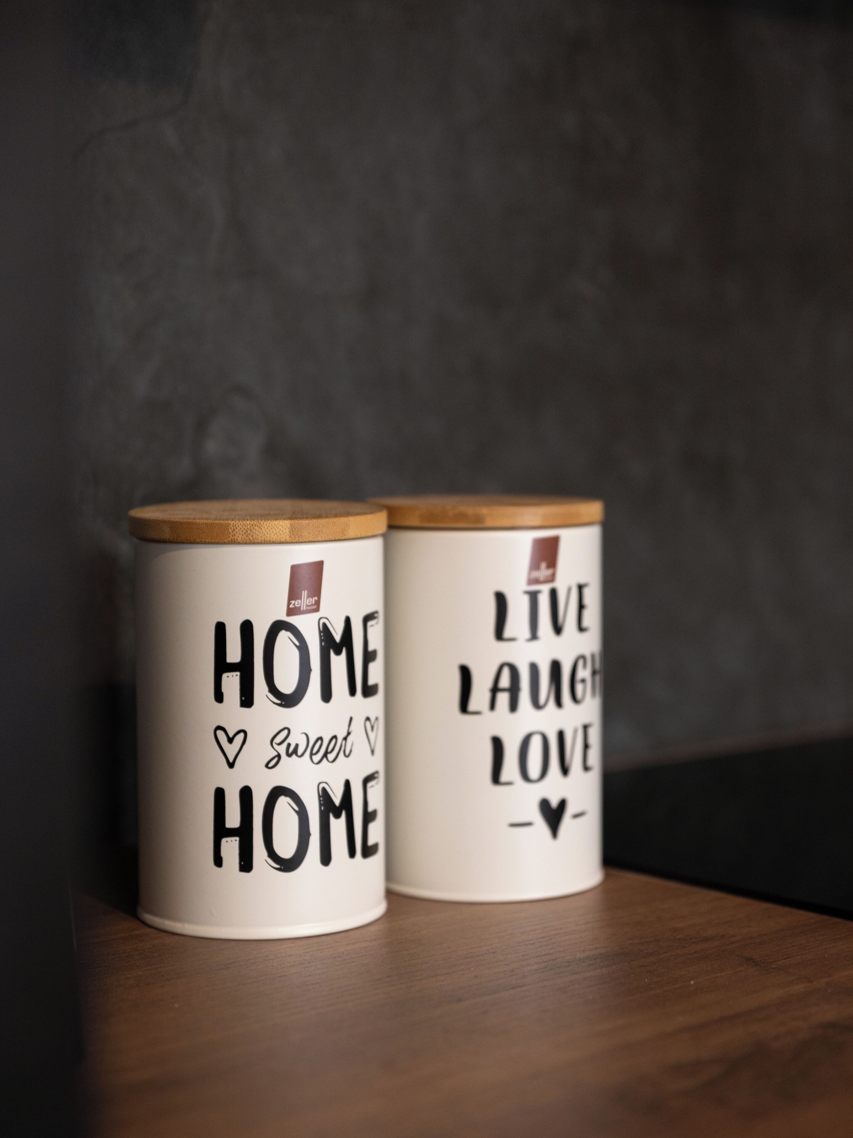 Due tazze di ceramica bianca con coperchi in legno, una con la scritta “HOME SWEET HOME” e l’altra con “LIVE LAUGH LOVE”.