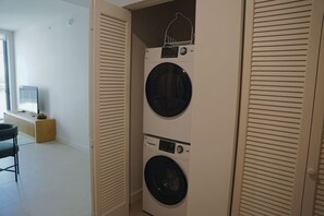 A laundry room with two stacked front-loading washing machines, a wooden bench, and a television in the background. - Stylish Minimalist 1BR in The Crosby (Miami)