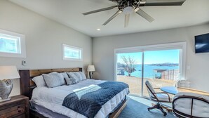 A bedroom with a large bed, a ceiling fan, a desk with a chair, and a view of the sea.