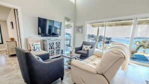 A modern living room with a large TV, comfortable seating, and a view of the sea.