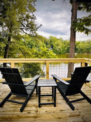 Terraza con dos sillas Adirondack negras y una pequeña mesa, con vista a un lago, árboles y un edificio distante.