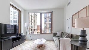Un salon moderne doté d’une grande fenêtre, d’un téléviseur à écran plat, d’un lampadaire, d’un canapé et d’une table basse.