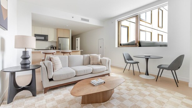 En moderne stue med sofa, sofabord og lænestole, et køkken i baggrunden og store vinduer.