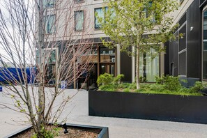 A modern building with a glass facade, a black planter with greenery, and a tree in the foreground. - Blueground | Greenpoint, pool, nr East River (Brooklyn)