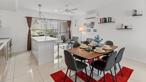 Cozinha moderna com ilha central, área de jantar com cadeiras e mesa para quatro, e sala de estar com sofá e abajur.