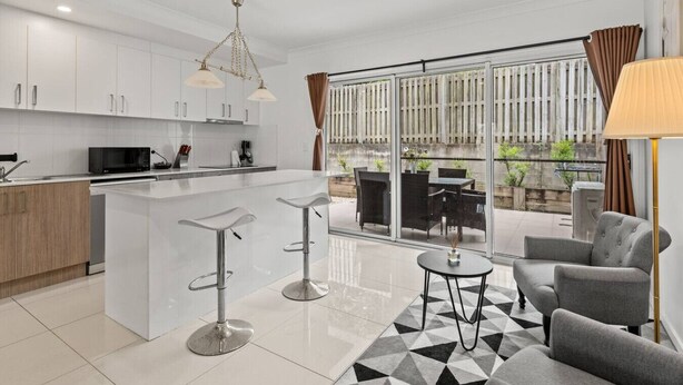 Cozinha moderna com armários brancos, uma ilha central com banquetas, sala de jantar com mesa e cadeiras, e uma grande porta de vidro deslizante que dá para uma varanda com plantas.