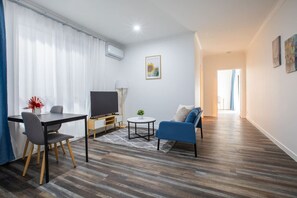 Una sala de estar moderna con un televisor de pantalla plana, una mesa de comedor con sillas, un sofá y una pequeña mesa redonda con una planta.