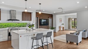 A modern kitchen with a central island, bar stools, a dining area with sofas, and a large window. - 3BR Family Home in Rochedale | Aussie Local Living (Rochedale)
