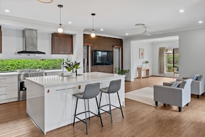 A modern kitchen with a central island, bar stools, a dining area with sofas, and a large window. - 3BR Family Home in Rochedale | Aussie Local Living (Rochedale)