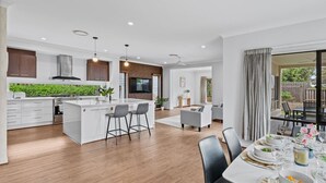 A modern kitchen with a central island, bar stools, a dining area with a table set for a meal, and a view of the outdoors. - 3BR Family Home in Rochedale | Aussie Local Living (Rochedale)