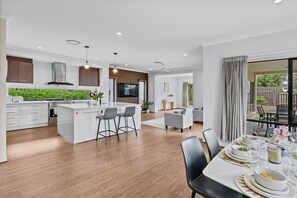 A modern kitchen with a central island, bar stools, a dining area with a table set for a meal, and a view of the outdoors. - 3BR Family Home in Rochedale | Aussie Local Living (Rochedale)