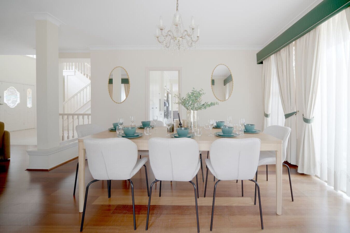 Un comedor con una mesa de madera preparada para cuatro personas, sillas blancas y una araña.