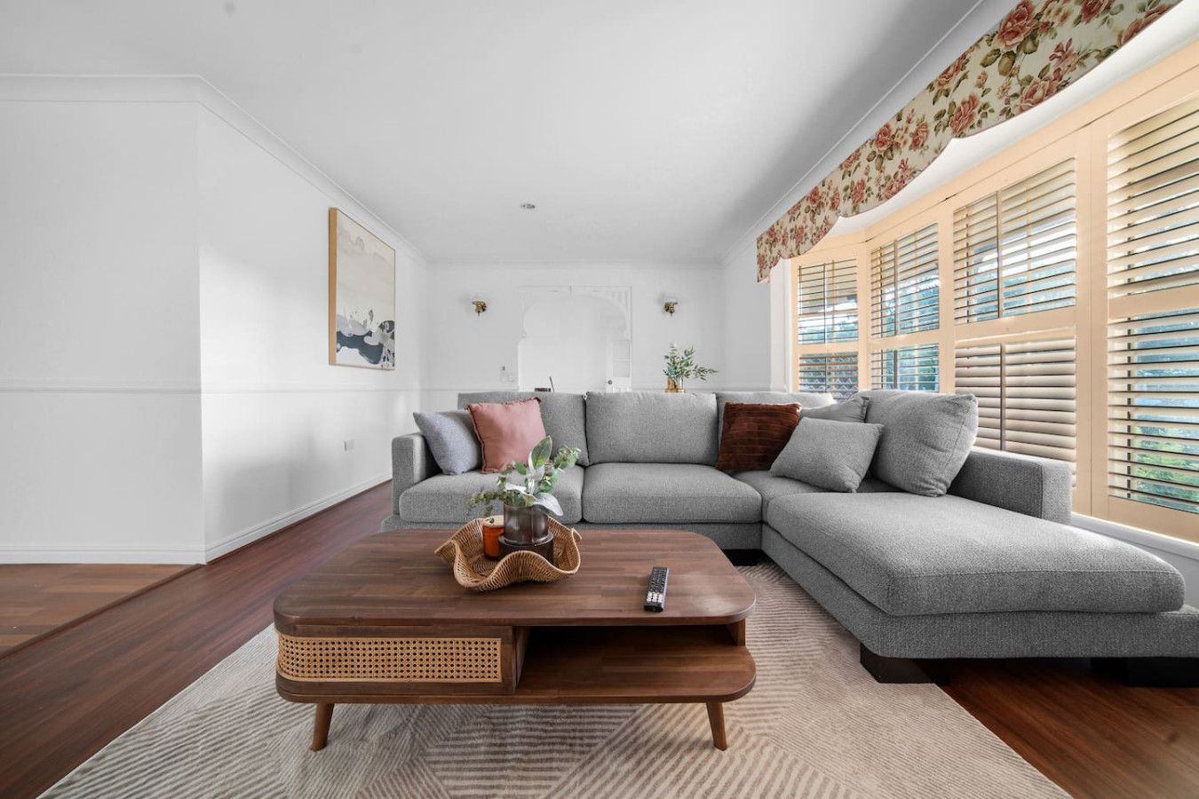 A modern living room with a grey sectional sofa, a wooden coffee table, and a large window with blinds.