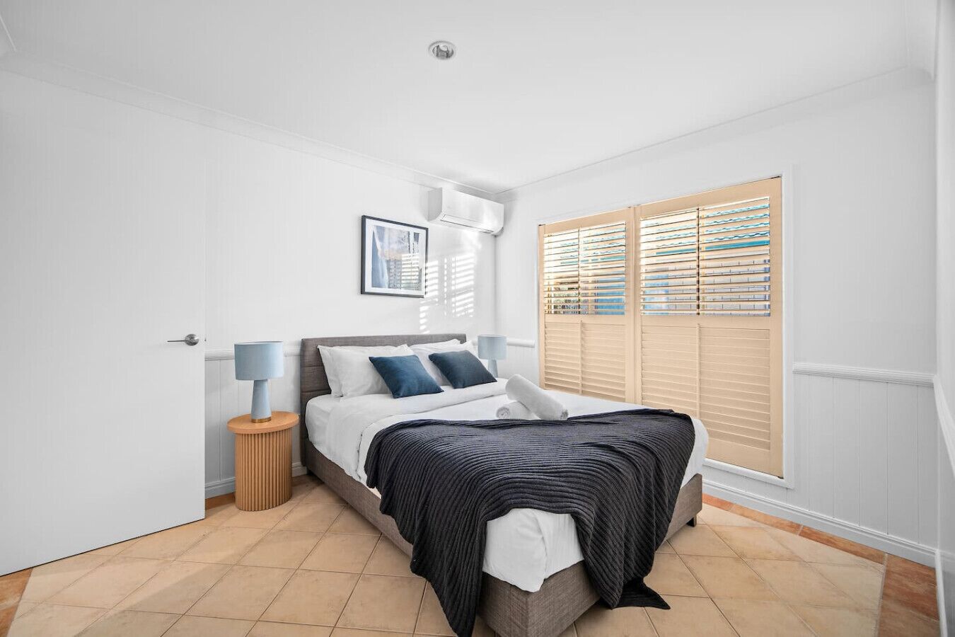 Une chambre à coucher comprenant un lit, des tables de chevet, une fenêtre avec des stores et une bouche d’aération au plafond.