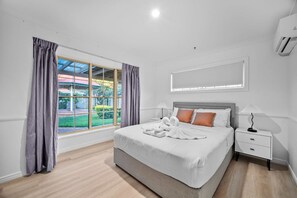 A hotel room with a large bed, a nightstand, a lamp, and a view of a green landscape through a window. - Modern 4B Family-Friendly Home in Robertson QLD (Robertson)