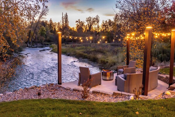 An outdoor seating area with a river view, surrounded by trees and string lights.