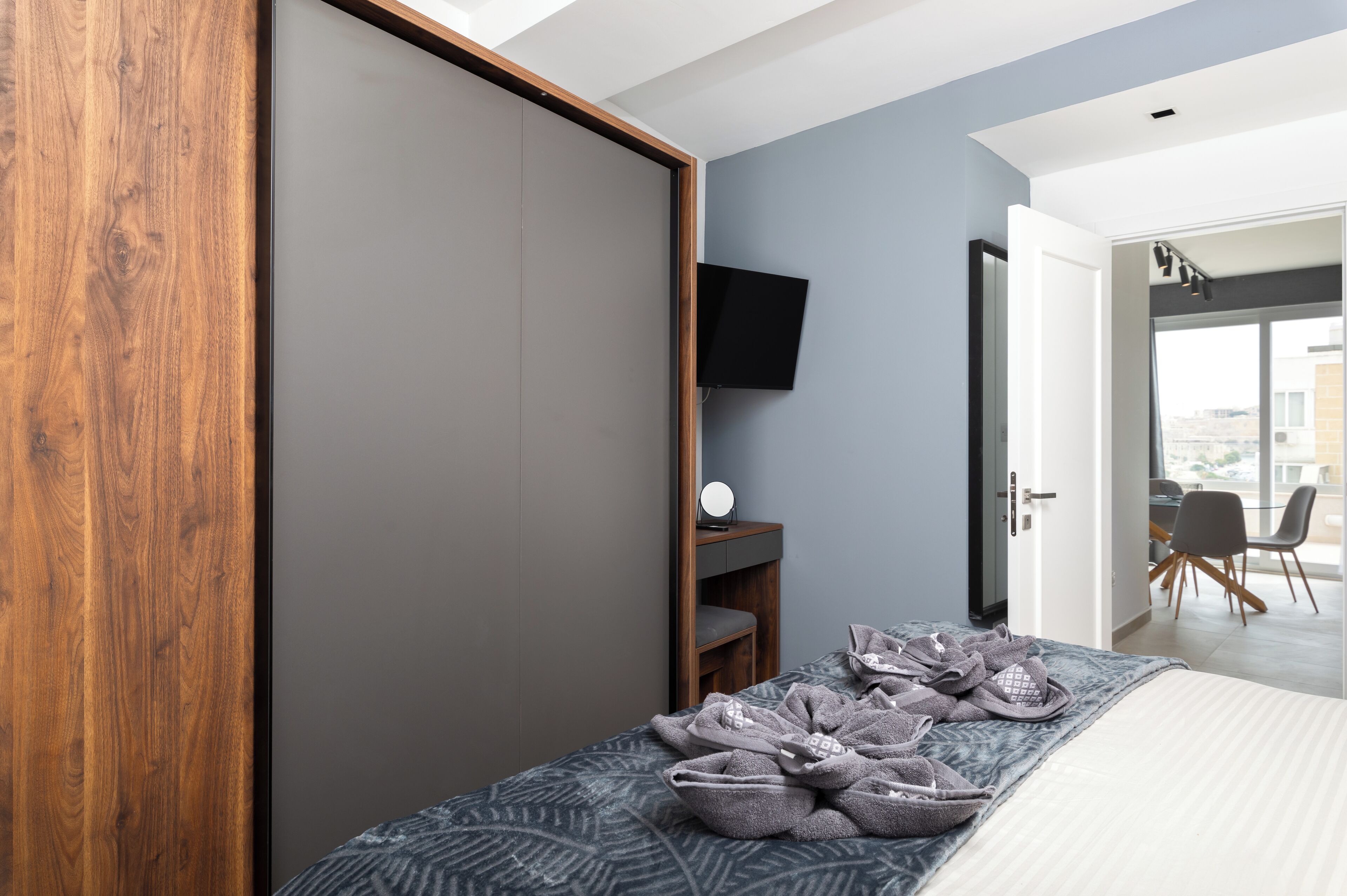 A modern hotel room with a large wardrobe, a flat-screen TV mounted on the wall, a small bedside table with a mirror, and a bed with a patterned blanket.