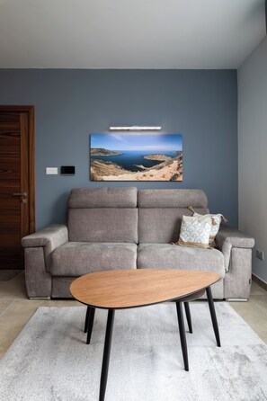 Uma sala de estar com um sofá cinza, uma mesa de centro de madeira e um quadro com uma paisagem na parede.