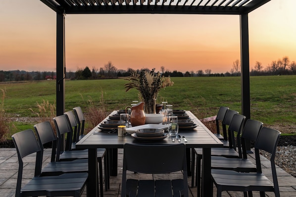 A rectangular outdoor dining table set for six with black chairs, surrounded by a pergola, set against a sunset view.