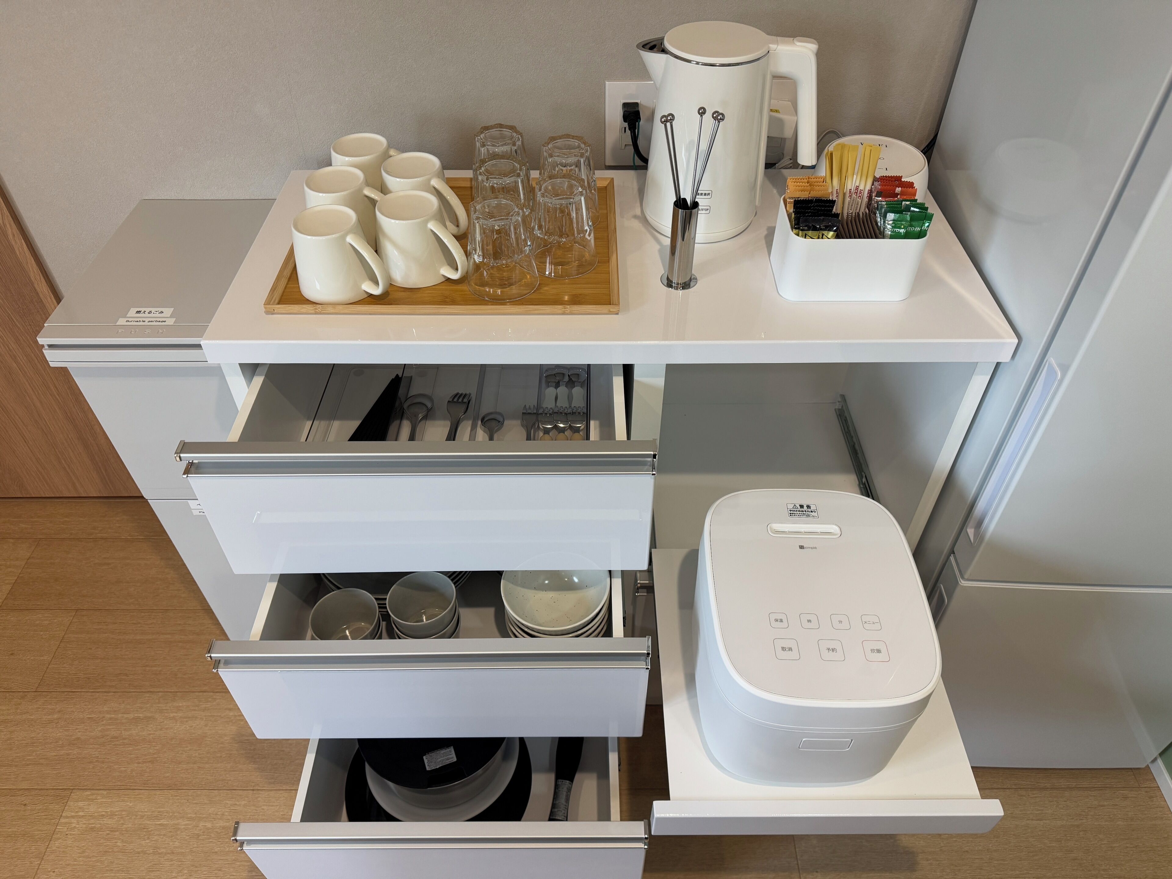 A kitchen area with a countertop holding mugs, glasses, a kettle, and utensils. There are drawers below with dishes and a rice cooker.