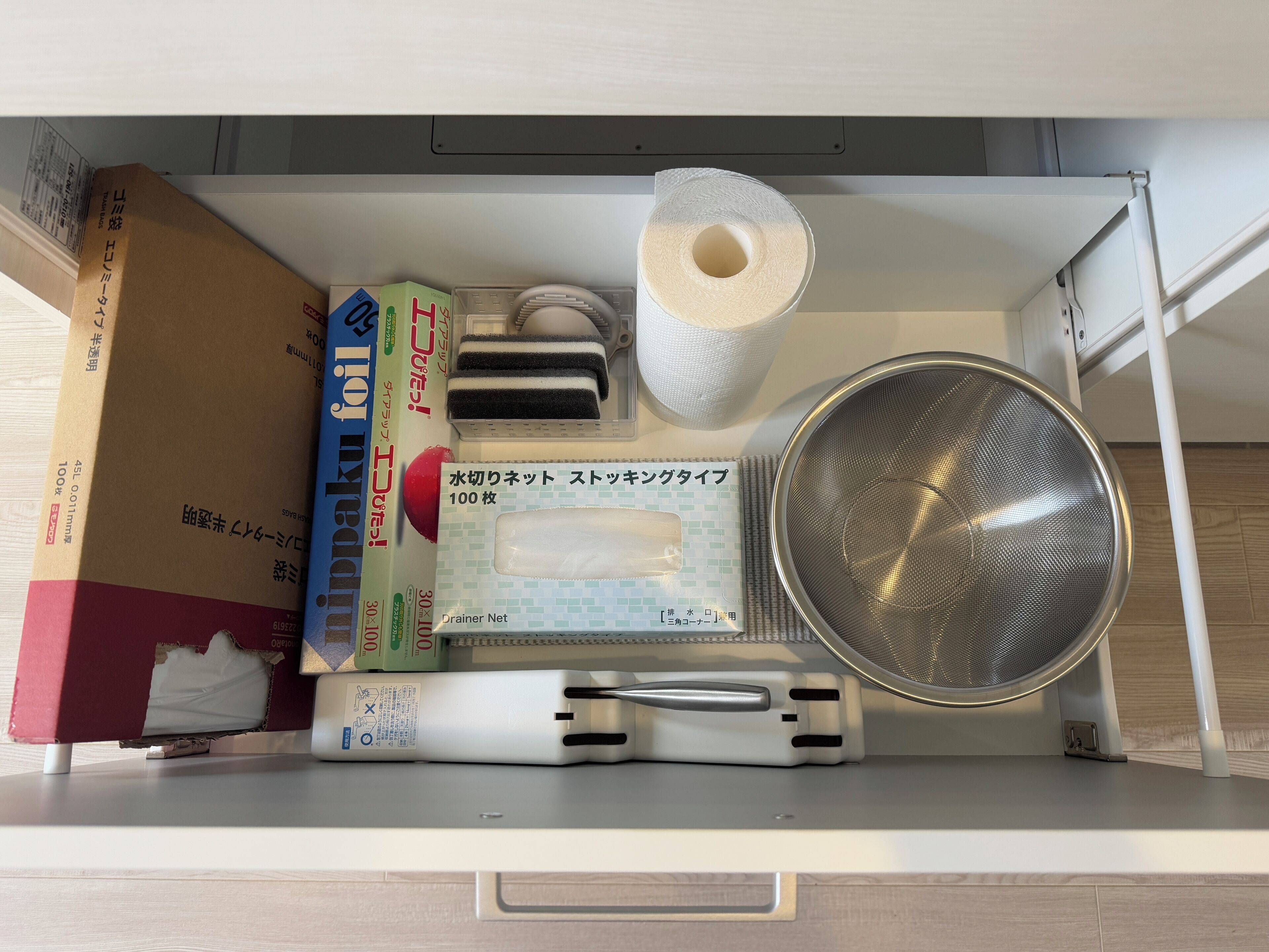 A drawer with various items including a box of toilet paper, a white plastic container, a metal strainer, and a white object with a handle.