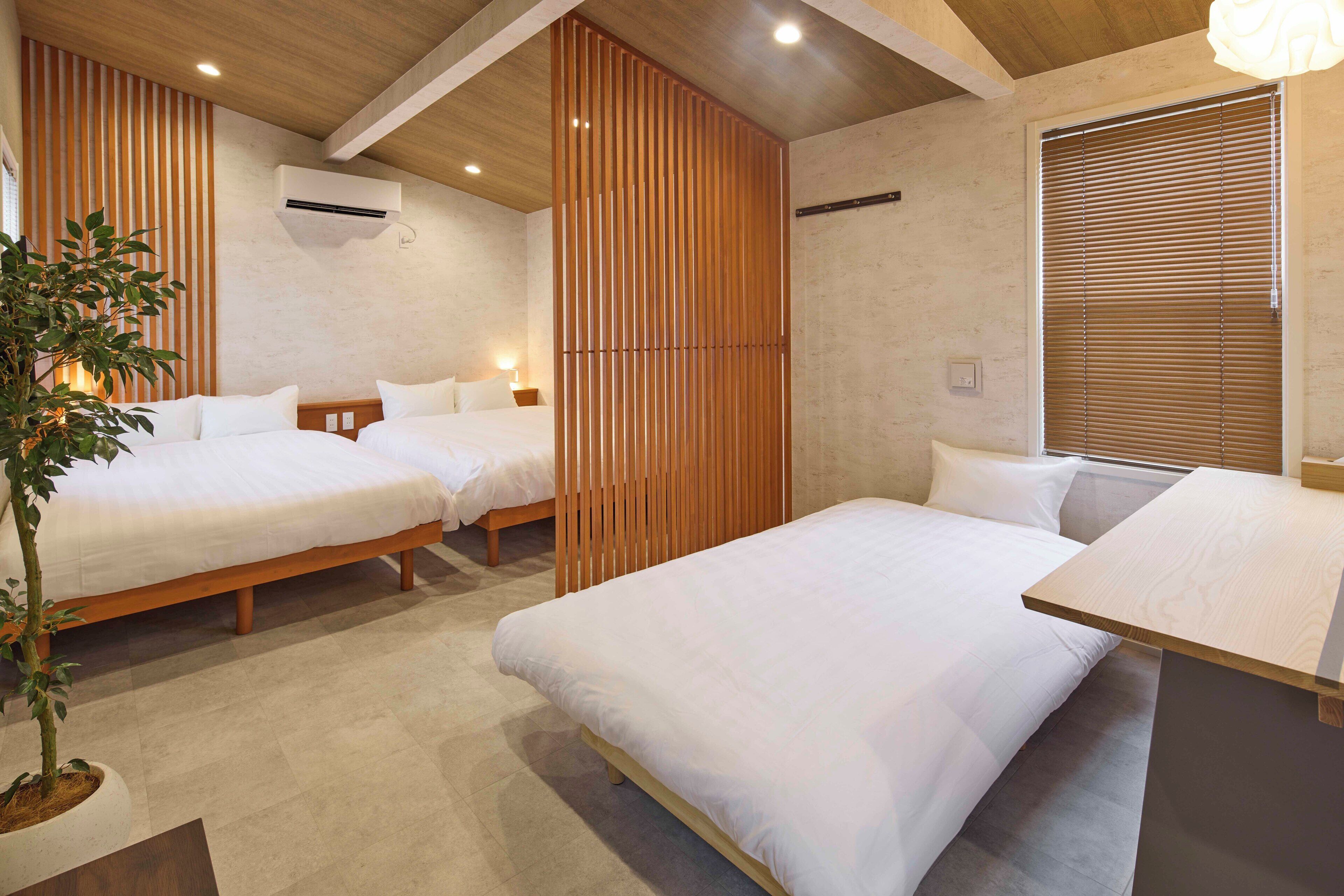 A modern hotel room with two beds, a wooden partition, a potted plant, and a window with blinds.