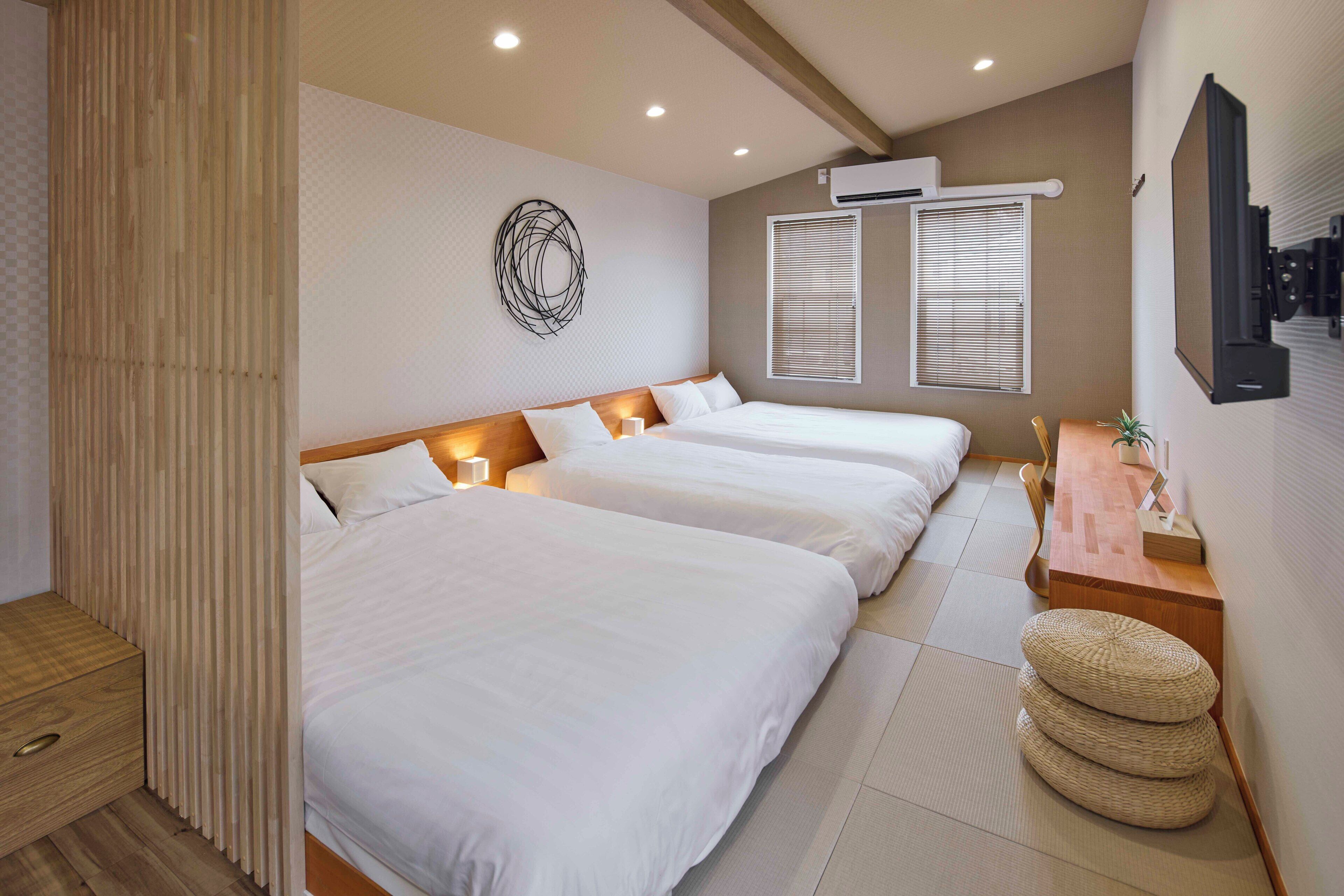 A hotel room with three beds, a wooden nightstand, a wall-mounted TV, and a window with blinds.