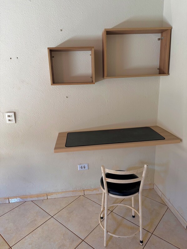 A corner room with a wooden desk, two wall-mounted shelves, a chair, and tiled flooring.