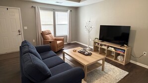 A living room with a dark blue sofa, a wooden coffee table, a TV stand with shelves, a ceiling fan, and a door with a window. - Tremendous 3-bedroom townhome with close to Cave Hollow Park in Warrensburg (Warrensburg)