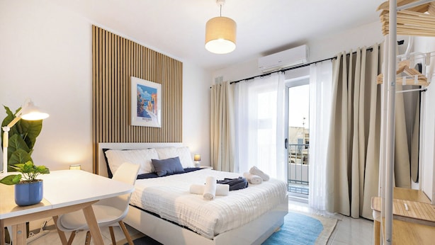 Habitación de hotel moderna con cama, escritorio, silla, planta en maceta, un cuadro en la pared y vistas al exterior.