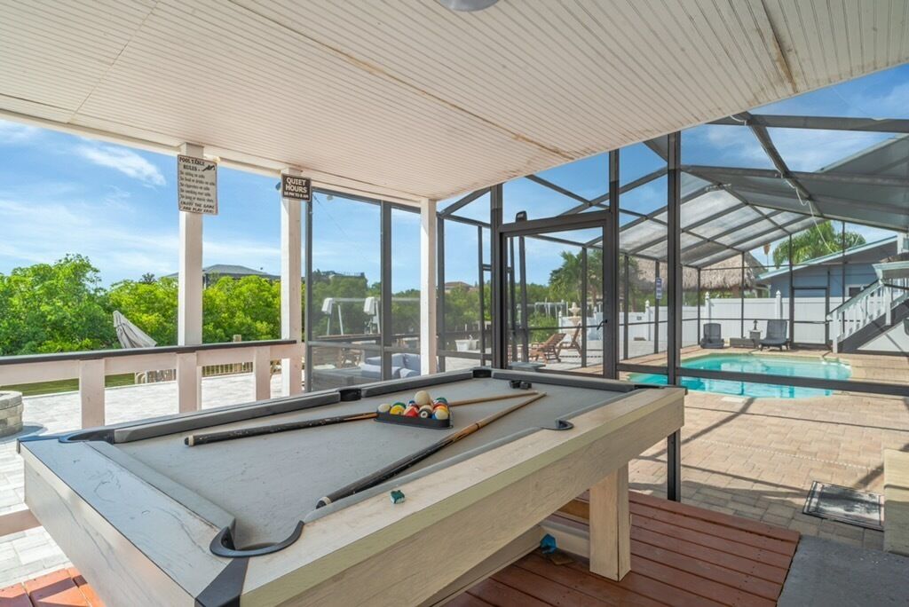 A pool table in a covered area with a view of a swimming pool and outdoor furniture.