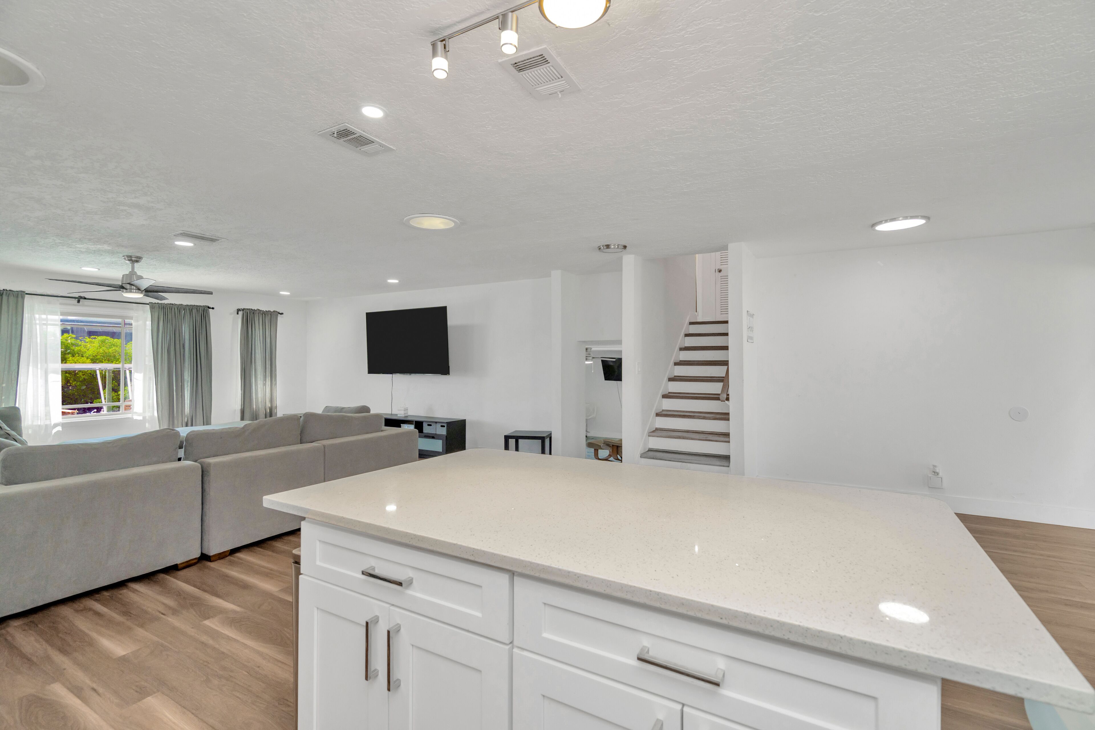 Un salon moderne avec un canapé d’angle gris, un îlot de cuisine blanc et un escalier menant à un niveau supérieur.