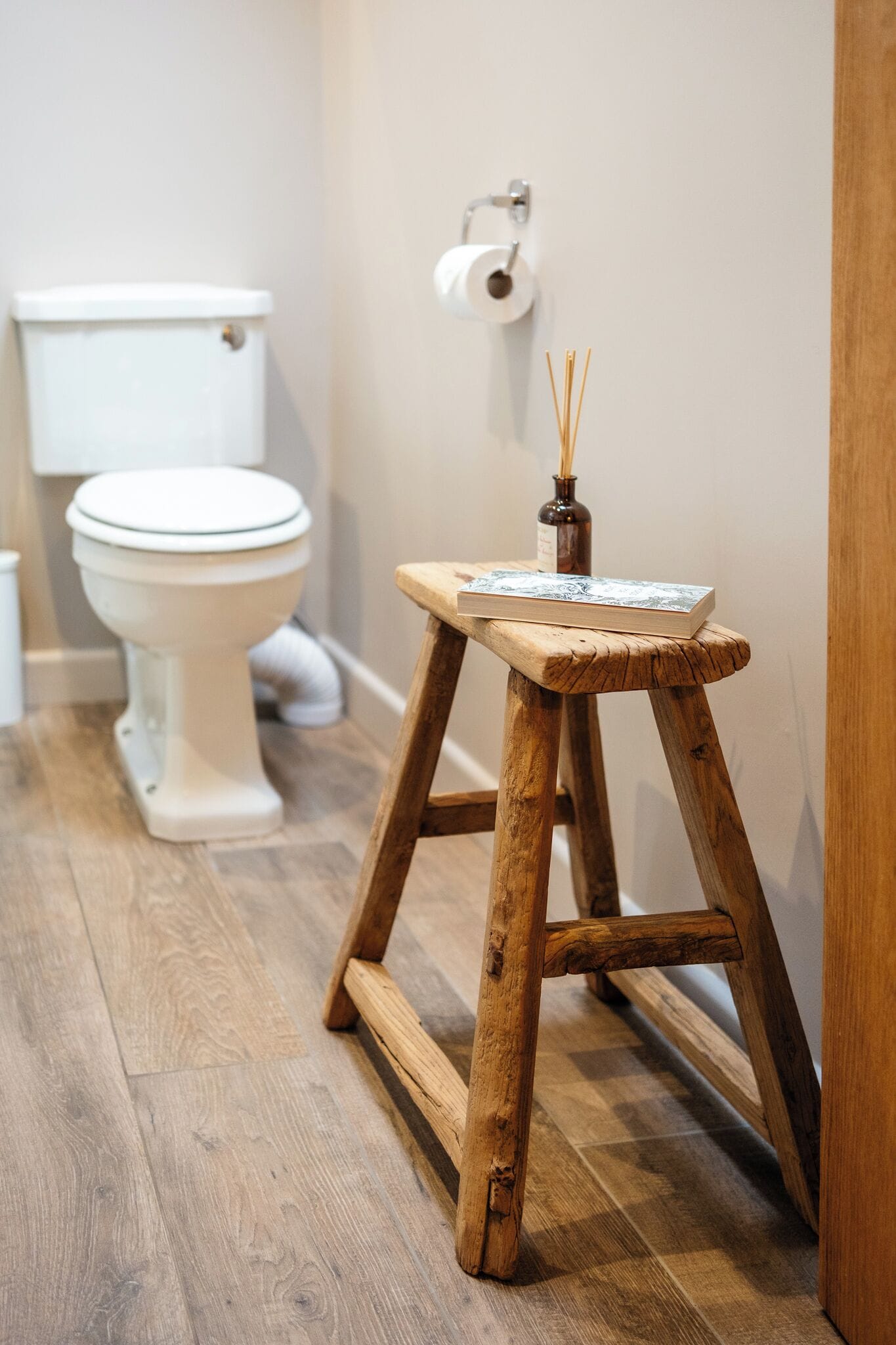 En træskammel med en bog og en rør diffusor i et badeværelse med et toilet og hvidt toiletpapir.
