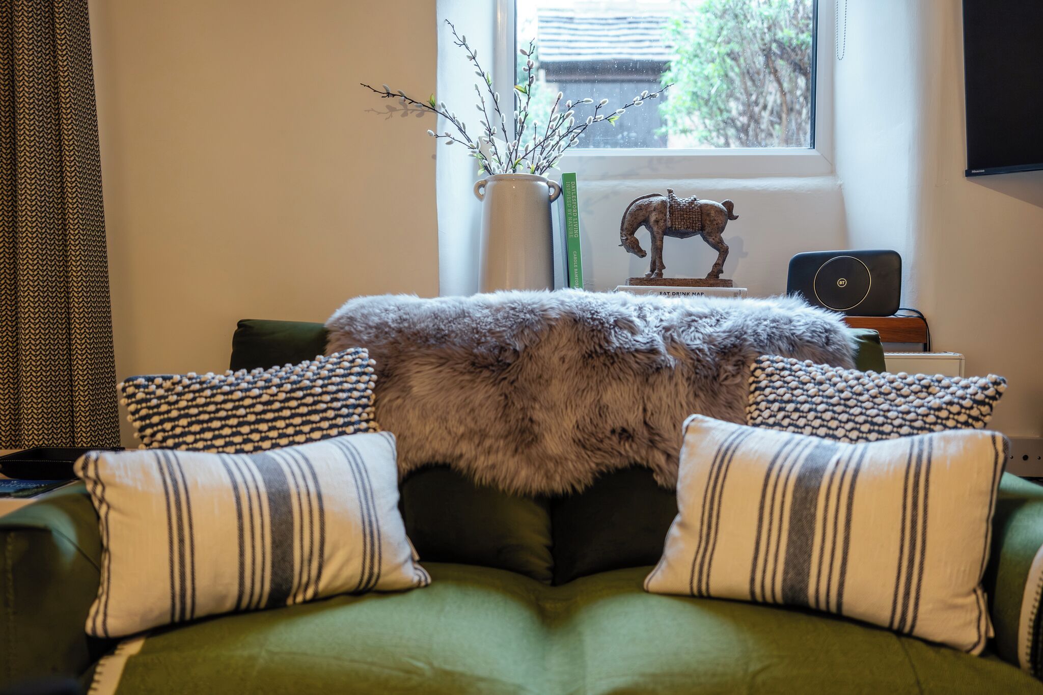A cozy living room with a green sofa, striped pillows, a fur throw, a small table with a decorative horse statue, a vase with branches, and a laptop on a side table.