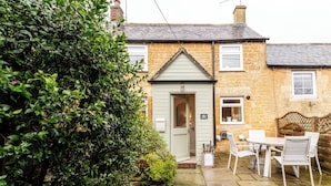 A traditional stone house with a small front garden, a white door, and a patio with a table and chairs. - Beautiful 2 BD Cottage in the Cotswolds (Moreton-in-Marsh)