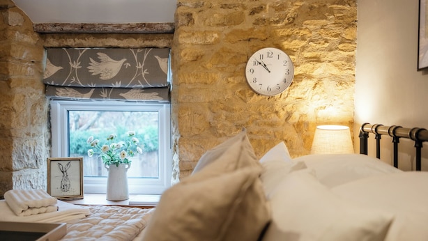 A bedroom with a stone wall, a clock, a window with a view, a bedside lamp, and a framed picture on the nightstand. - Beautiful 2 BD Cottage in the Cotswolds (Moreton-in-Marsh)