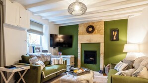 A cozy living room with green sofas, a flat-screen TV, a fireplace, and a modern chandelier. - Beautiful 2 BD Cottage in the Cotswolds (Moreton-in-Marsh)