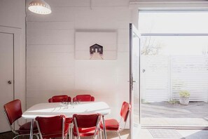 A dining area with a white table and red chairs, a framed picture on the wall, and a view of a white fence outside. - Ruby SoHo Workshop Villa- Port Sorell (Latrobe)