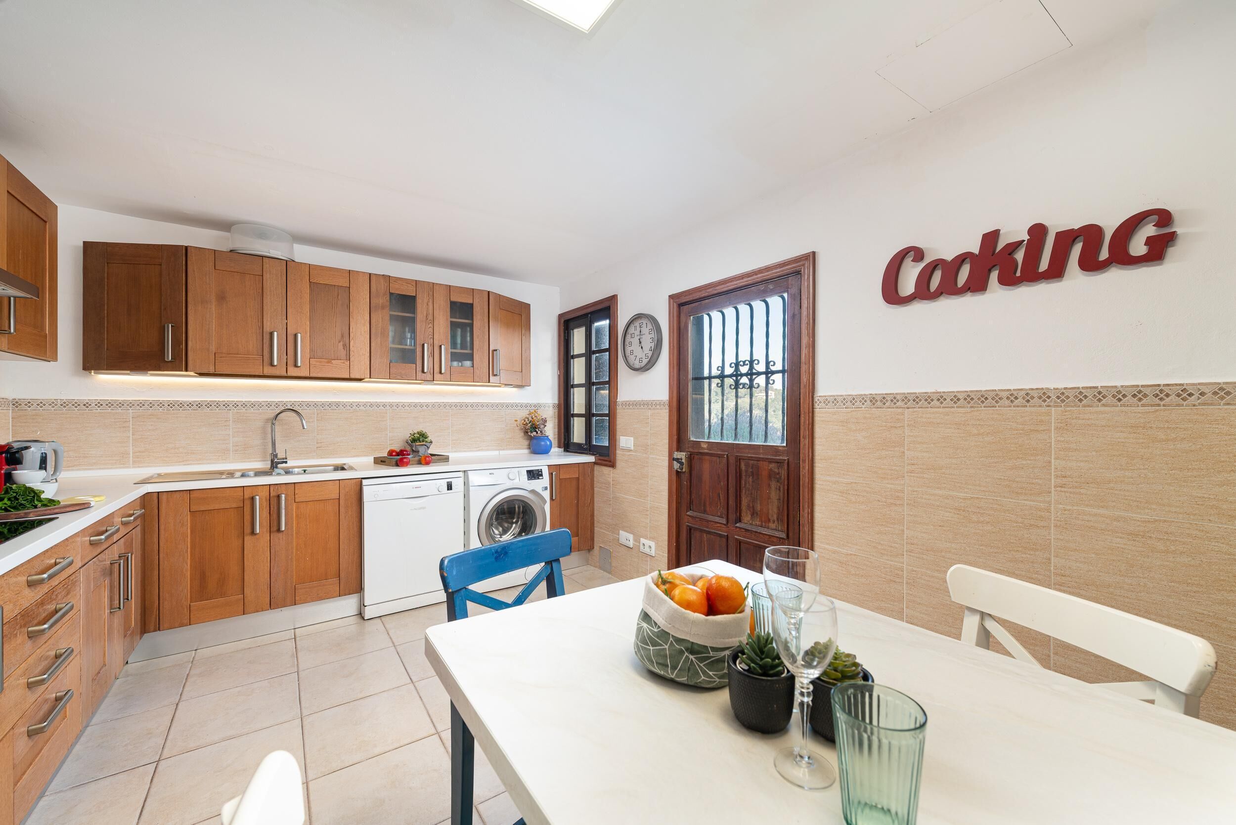 A kitchen with wooden cabinets, a washing machine, a sink, and a table with a bowl of fruit and glasses.