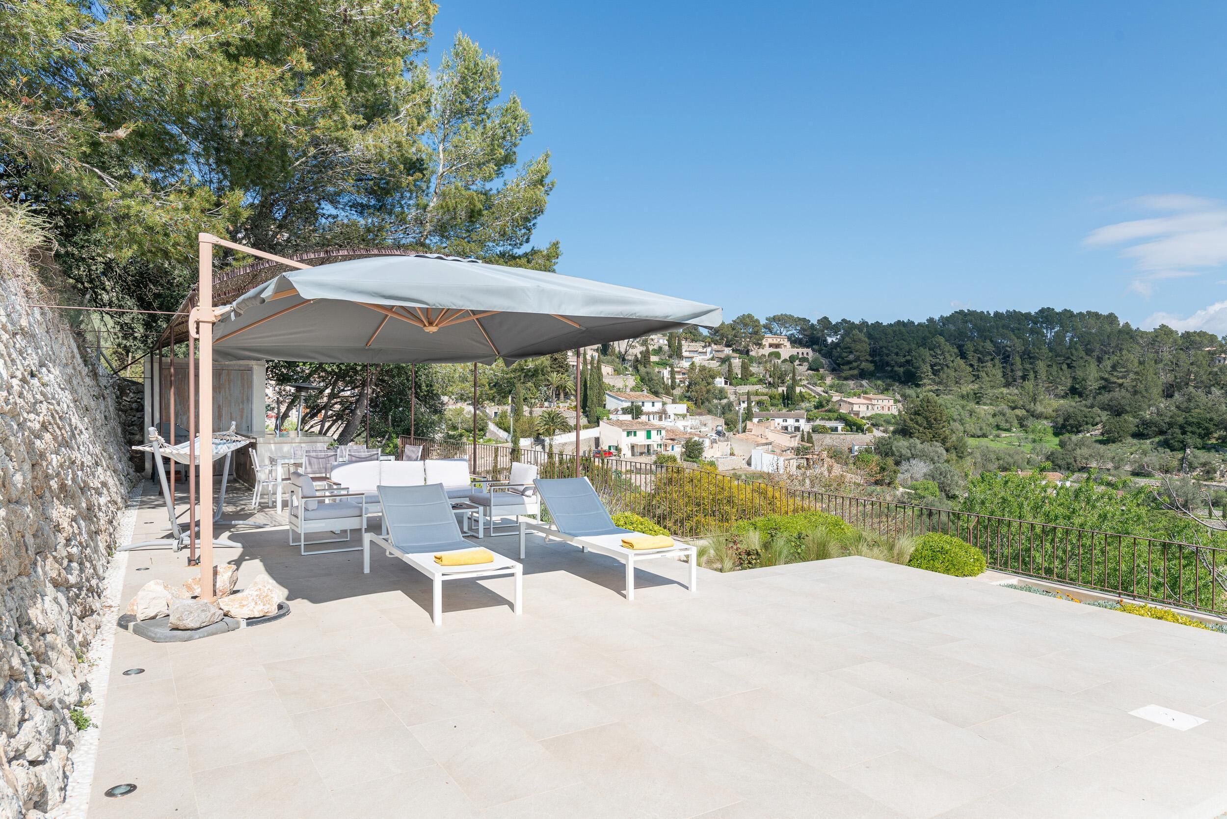 An outdoor seating area with tables and chairs, a covered umbrella, and a view of a hillside town.