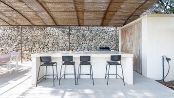 Un área de bar al aire libre con un fondo de pared de piedra, techo de paja y mostrador de piedra con taburetes.