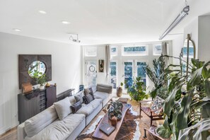 A spacious living room with a large sofa, a coffee table, and various plants. There is a large window providing natural light and a view of the outdoors. - Bohome Reserve:  Chicago Wicker Park • Sauna • Chef's Kitchen • Outdoor Lounging (Chicago)