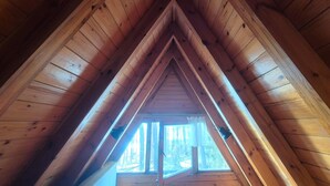 Une lucarne triangulaire avec des poutres en bois, offrant une vue sur les arbres à l’extérieur.