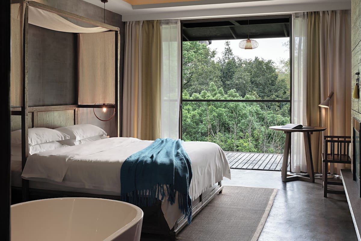 Un dormitorio con una cama grande, vistas al verdor exterior, una mesita pequeña y una silla.