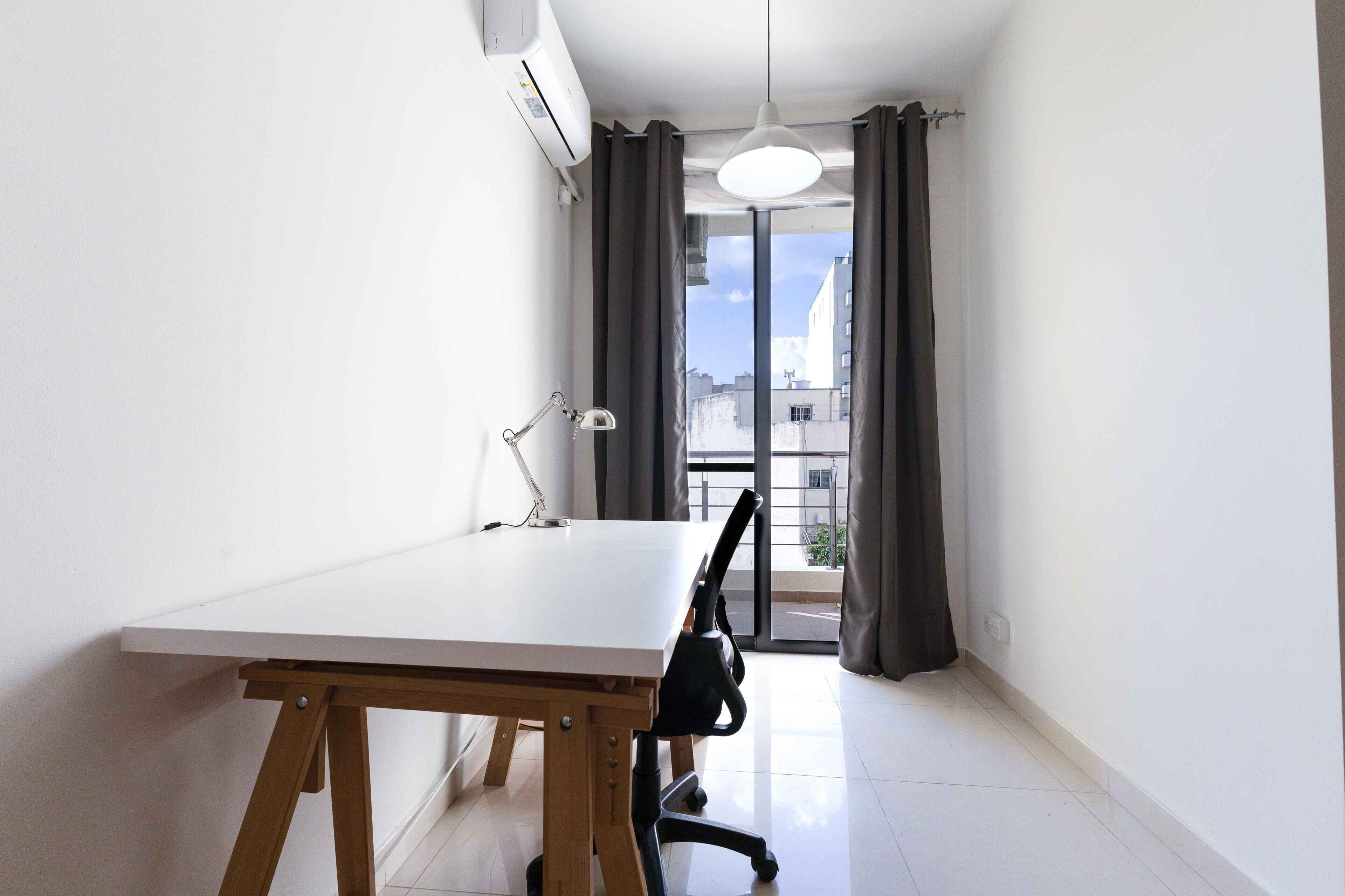 A small, bright room with a white table and a black office chair, a white desk lamp, and a window with curtains.