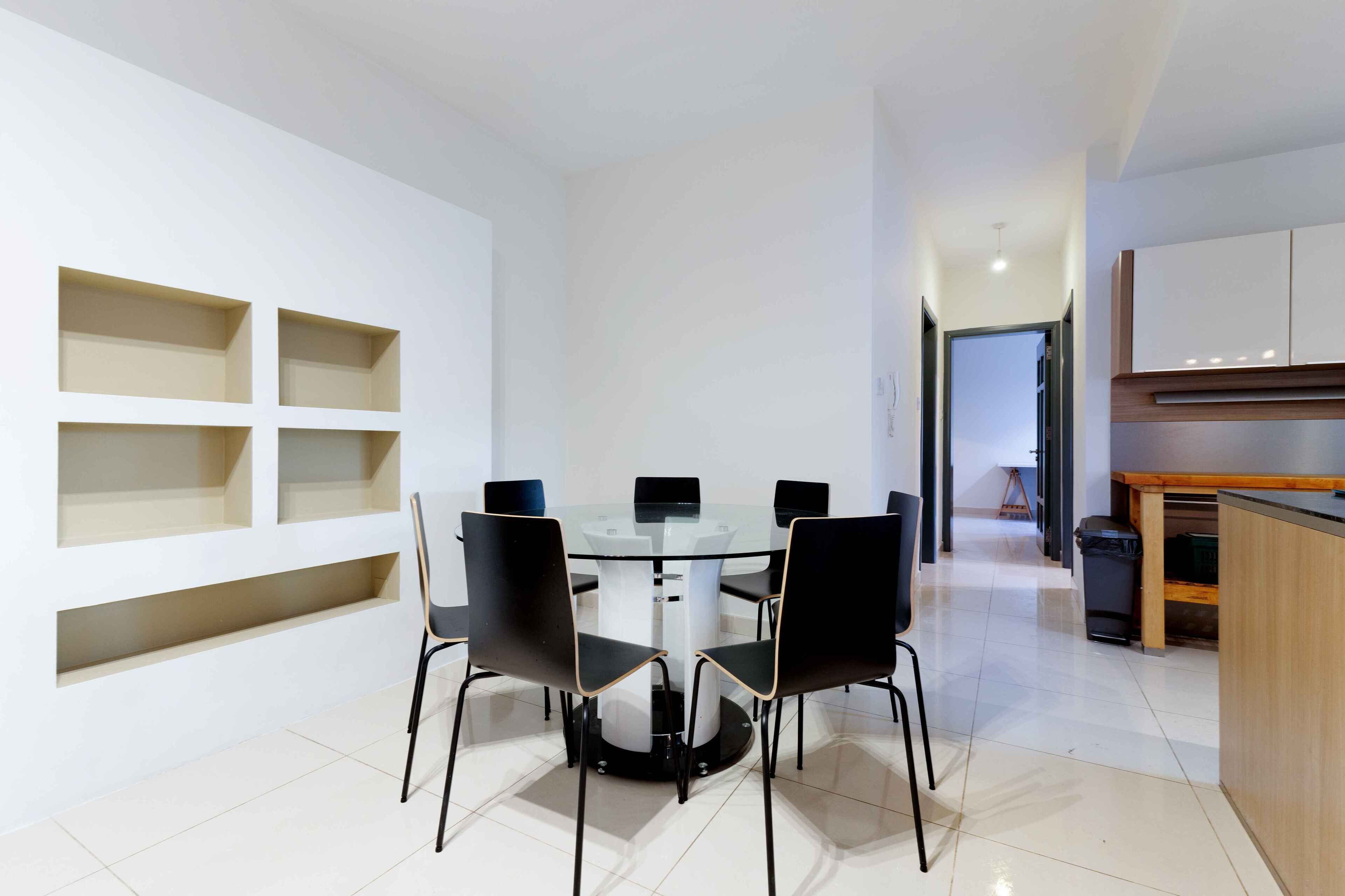 A modern dining area with a glass table surrounded by four black chairs, white walls with built-in shelves, and a kitchen area with wooden cabinets.
