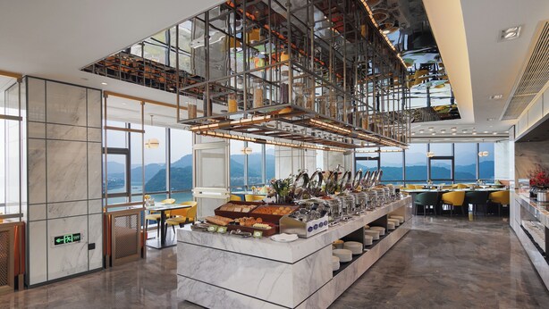 A modern hotel lobby with a marble reception desk, large windows offering mountain views, and a high ceiling with exposed structural elements.