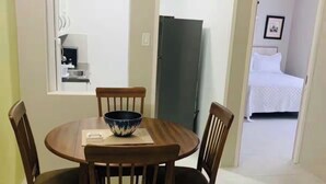 Un comedor con una mesa redonda de madera, cuatro sillas y un cuenco sobre la mesa. Hay una ventana sobre la mesa y una puerta a la derecha.